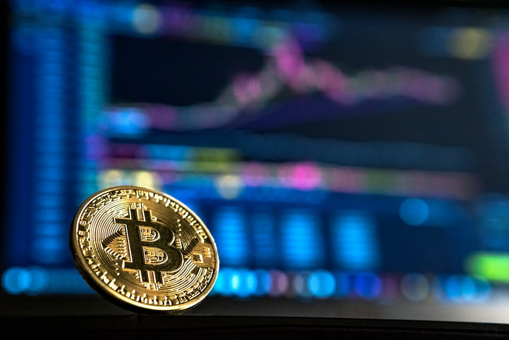 A Bitcoin coin on a table in front of a computer screen displaying a financial chart.