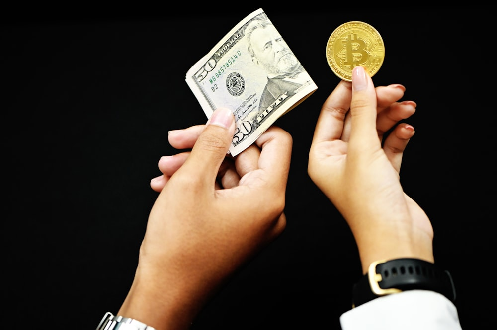 One hand holding a fifty-dollar bill and another hand holding a gold Bitcoin.