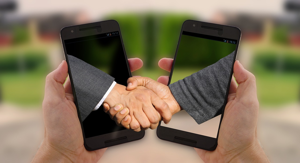 Two hands emerging from smartphone screens shaking hands.