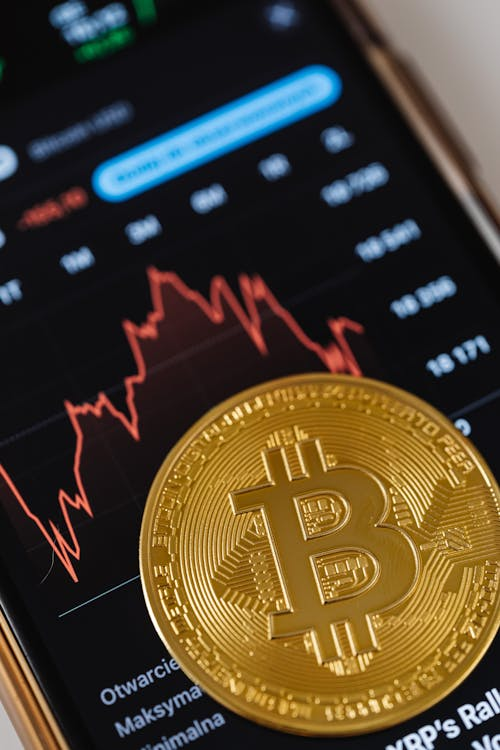 Close-up photo of a Bitcoin cryptocurrency coin resting on a smartphone screen displaying a financial graph.