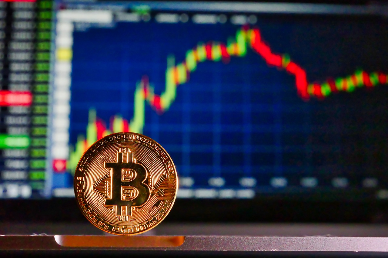 A close-up of a Bitcoin coin, with a laptop computer screen displaying a stock chart.