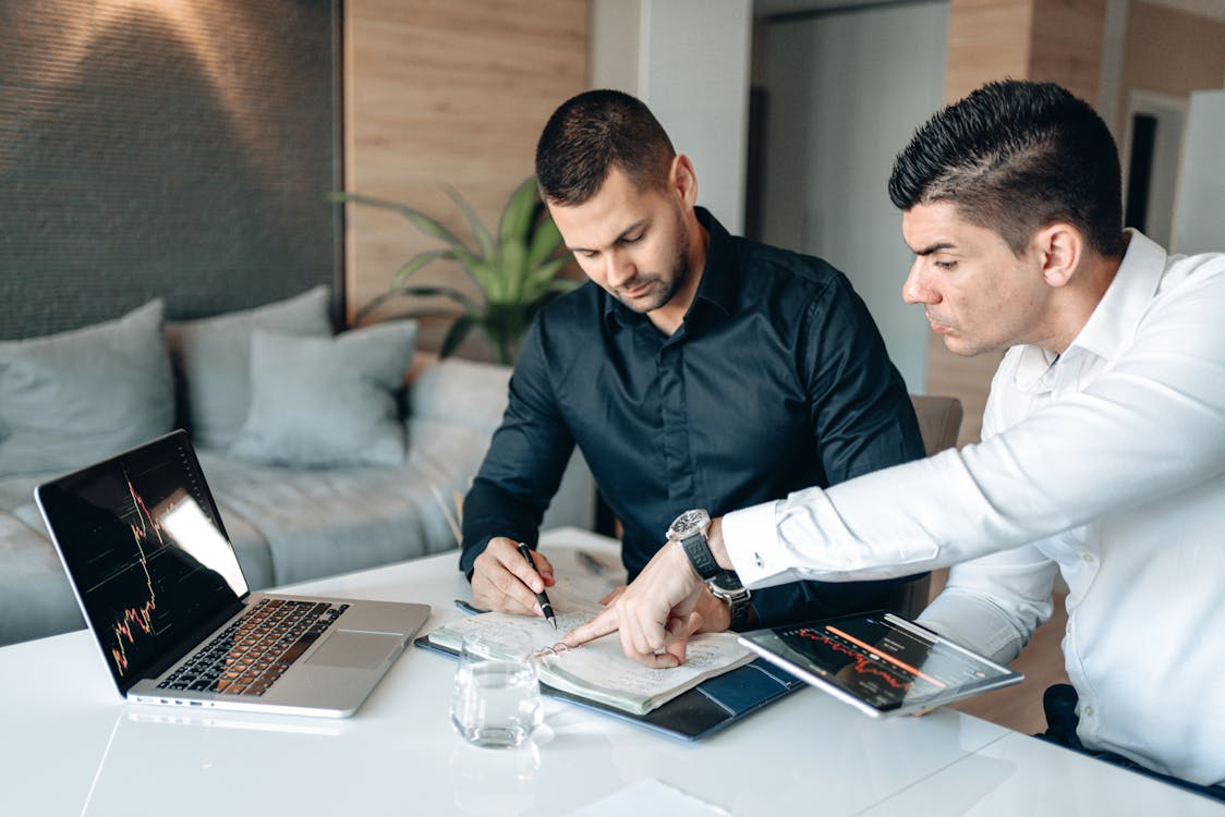 Two men discussing digital asset investment solutions with a laptop and a tablet displaying market data on a table.