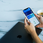 A person using a financial app on their smartphone beside a MacBook and coffee cup.