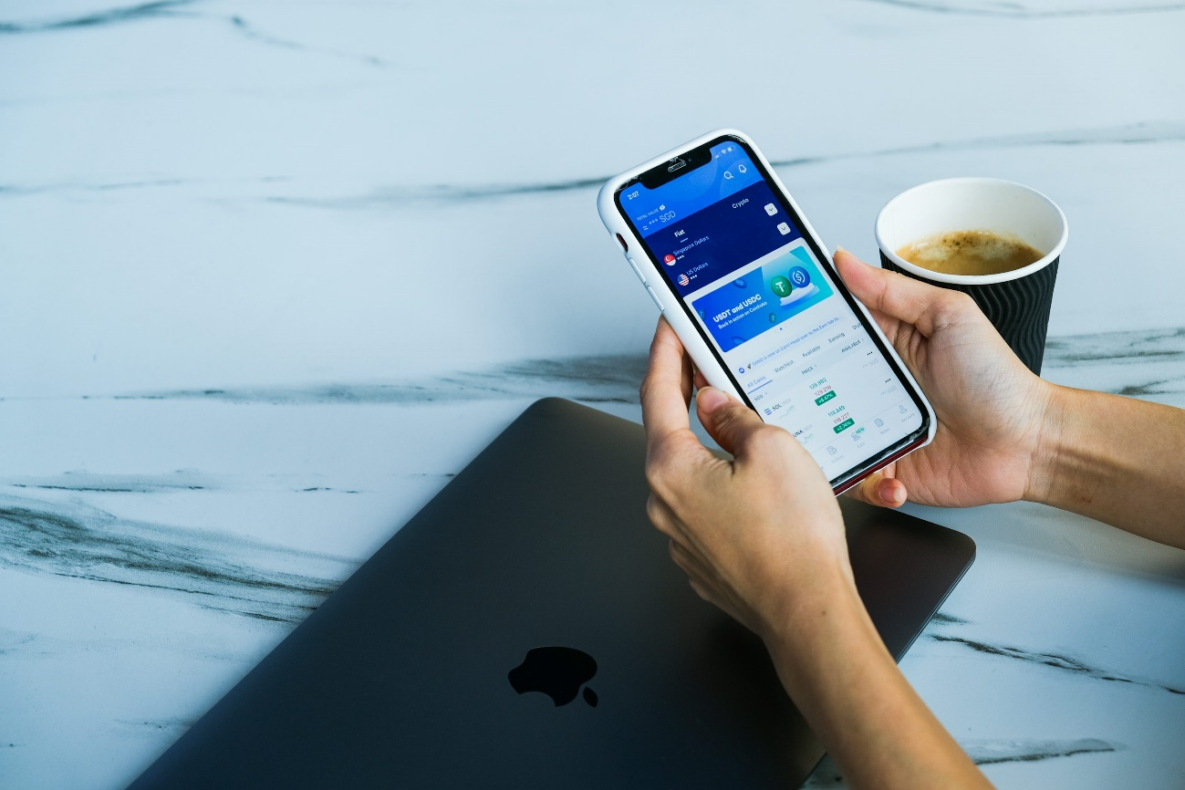 A person using a financial app on their smartphone beside a MacBook and coffee cup.