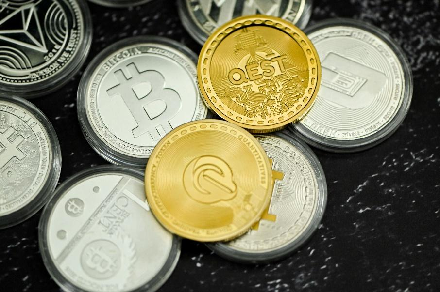 A close-up photo of various cryptocurrency coins, including Bitcoin, Quest, and Cent, displayed on a black surface.