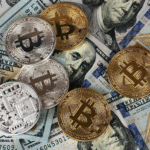 Several gold and silver-colored physical Bitcoin tokens scattered over multiple U.S. one hundred dollar bills.