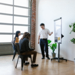 A man in a white shirt standing next to a flip chart, presenting to two seated individuals in a room with large windows and potted plants.