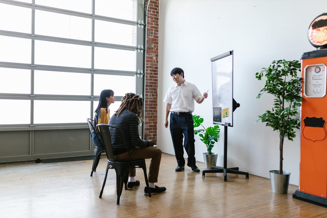 kenson Investments | When a 70% Drop Isn’t the Whole Story: The SharpLink Gaming Shake-Up A man in a white shirt standing next to a flip chart, presenting to two seated individuals in a room with large windows and potted plants.