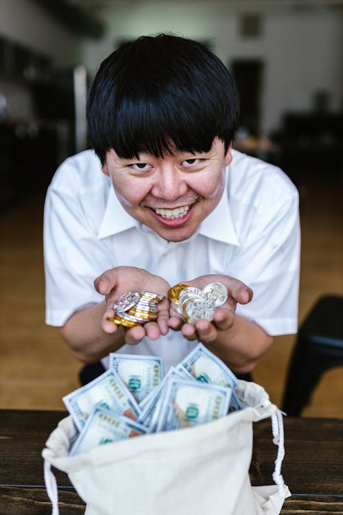 A smiling man in a white shirt holding stacks of silver and gold-colored physical cryptocurrency tokens in both hands.