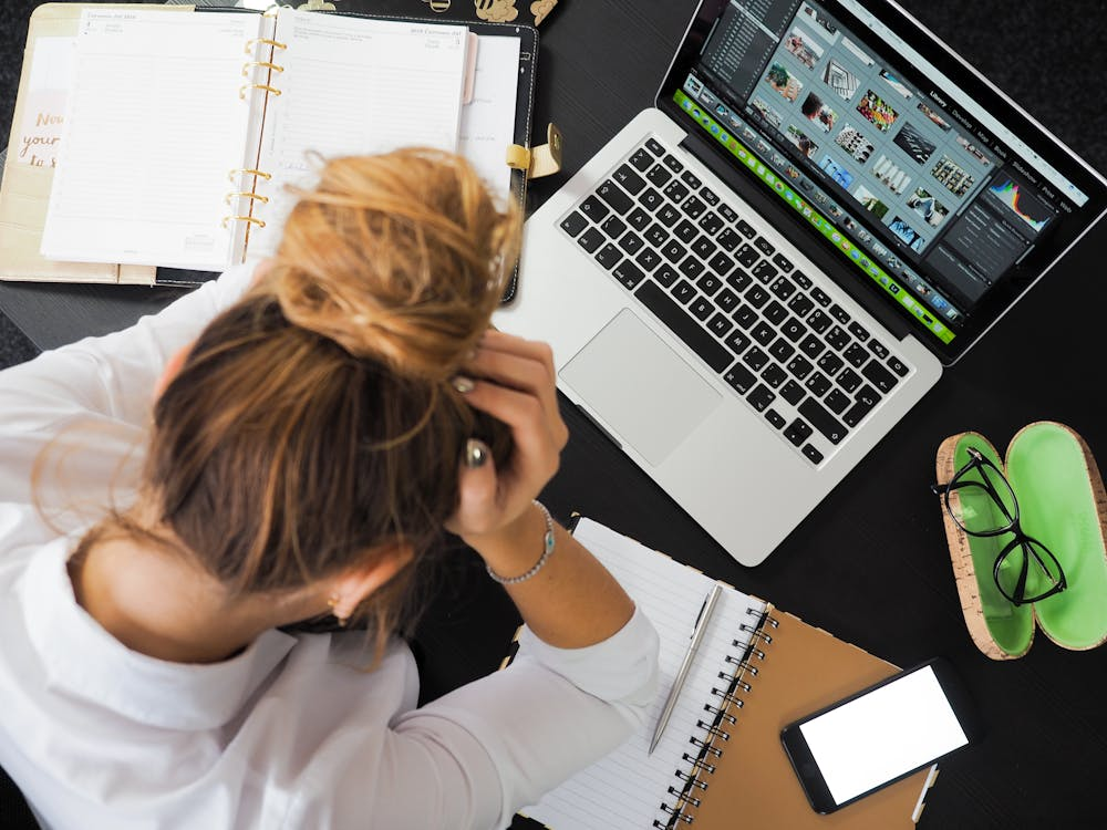 kenson Investments | Whales Are Surfacing: Big ETH Moves Amid Retail Retreat A woman with her hair in a bun sitting at a desk with her hands on her head, surrounded by a laptop, notebooks, smartphone, and eyeglasses.