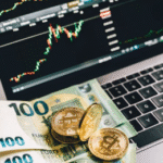 Gold-colored Bitcoin tokens and several 100 Euro banknotes placed on a laptop keyboard with trading charts displayed on the screen.