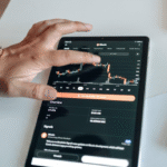 A person using a tablet displaying a candlestick chart of a cryptocurrency trading app, with one hand touching the screen.