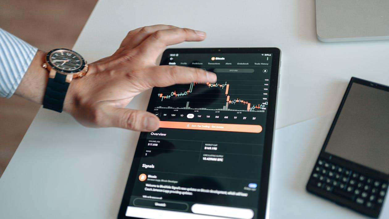 kenson Investments | Pompliano Goes Big: $750M Bitcoin Play Signals Institutional Boldness A person using a tablet displaying a candlestick chart of a cryptocurrency trading app, with one hand touching the screen.