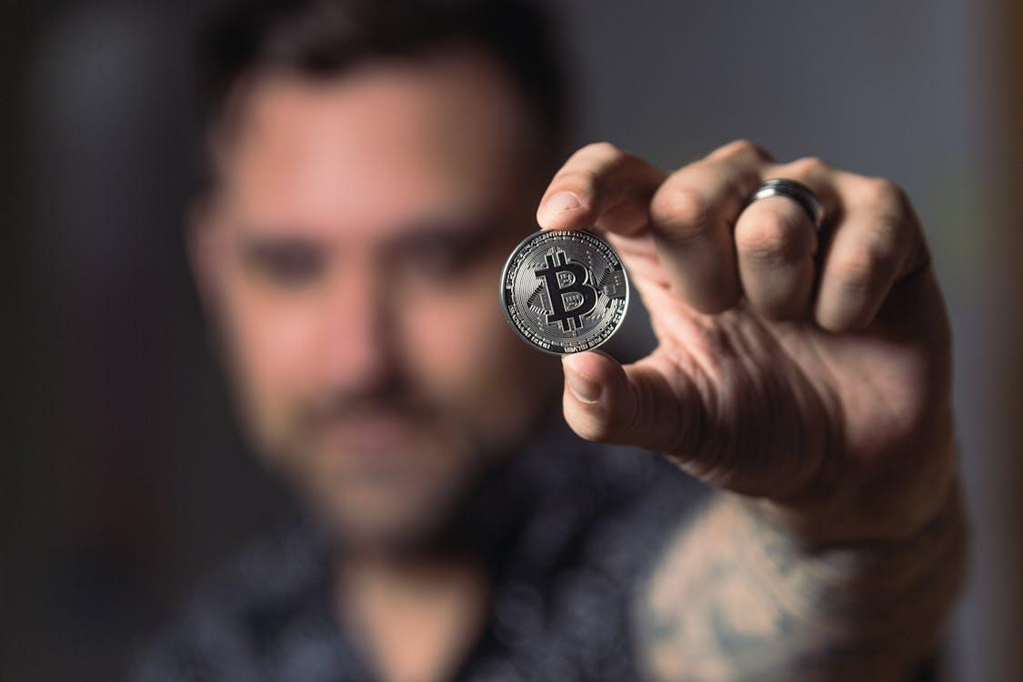kenson Investments | Pompliano Goes Big: $750M Bitcoin Play Signals Institutional Boldness A man holding a silver-colored physical Bitcoin token between his fingers with the focus on the coin and his face blurred in the background.