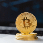 A stack of gold-colored physical Bitcoin tokens placed on a white surface in front of a blurred laptop keyboard.