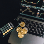 Gold and silver-colored Bitcoin tokens placed on a laptop keyboard next to a smartphone displaying a calculator app and a trading chart on the screen.