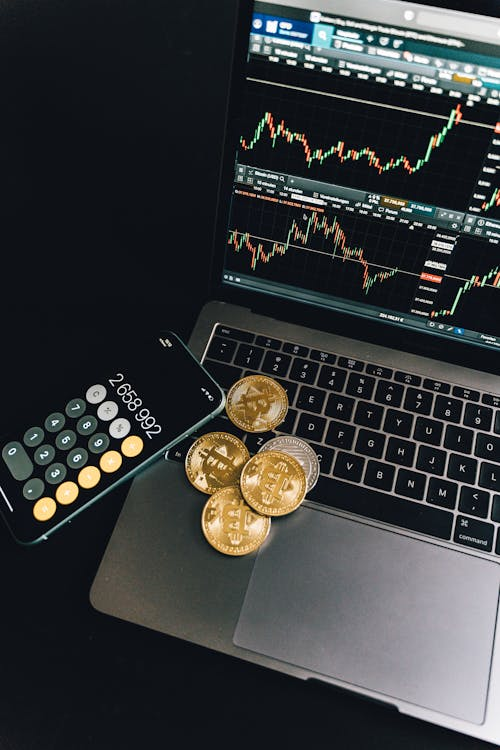 kenson Investments | Paul Tudor Jones Just Dropped a Bitcoin Bomb—And We’re Listening Gold and silver-colored Bitcoin tokens placed on a laptop keyboard next to a smartphone displaying a calculator app and a trading chart on the screen.