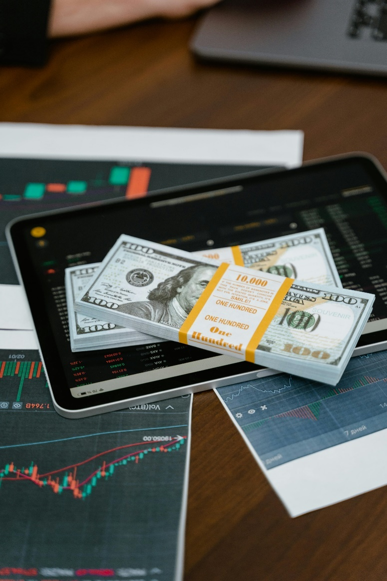 kenson Investments | Tokenization in 2025: Moving Beyond Real Estate and Into Private Credit and Infrastructure A tablet displaying financial charts and graphs, with stacks of US dollar bills.