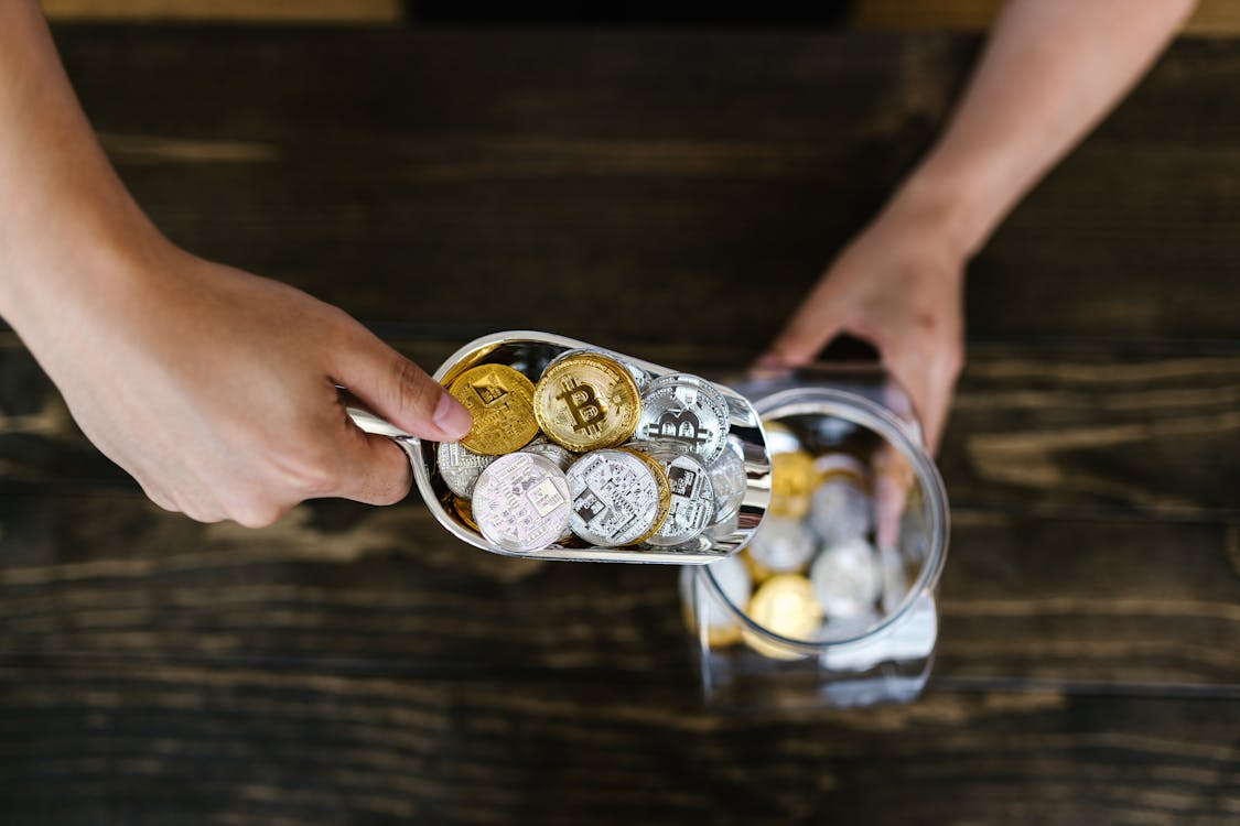 kenson Investments | Dogecoin Dips After the Hype – ETF Talk Isn’t Enough to Keep the Rally Going A person using a metal scoop to transfer silver and gold-colored physical cryptocurrency tokens into a glass jar on a wooden surface.