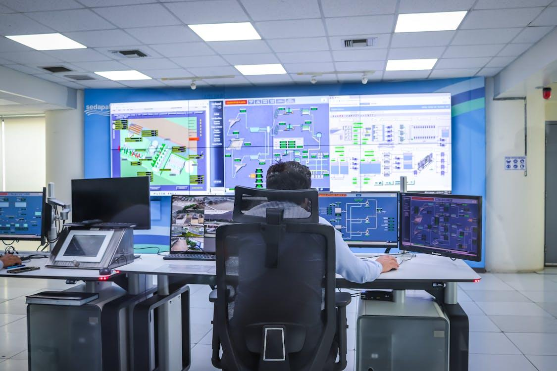 kenson Investments | Retail Meets Digital: Walmart and Amazon Eye Stablecoins in the U.S. A person sitting in a control room in front of multiple computer monitors and large wall screens displaying system diagrams and data dashboards.