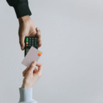 A person holding a pink credit card near a wireless card reader being held by another person, against a plain gray background.