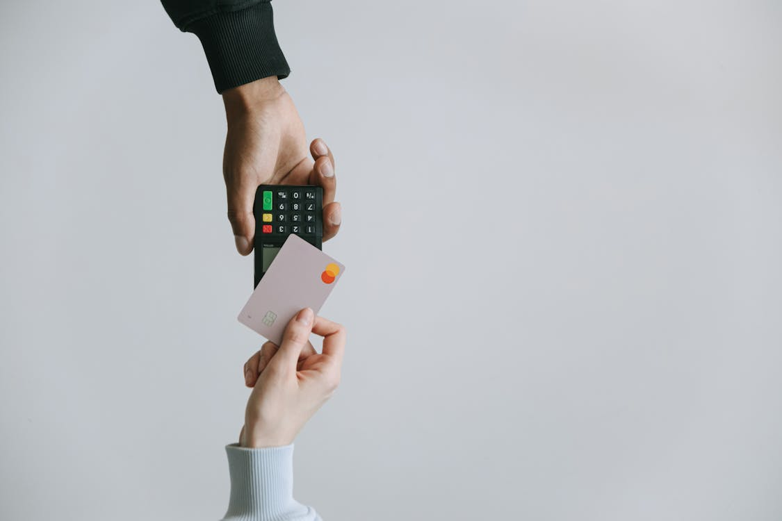 kenson Investments | Is XRP Really Gunning for SWIFT’s Turf? Let’s Talk Numbers A person holding a pink credit card near a wireless card reader being held by another person, against a plain gray background.