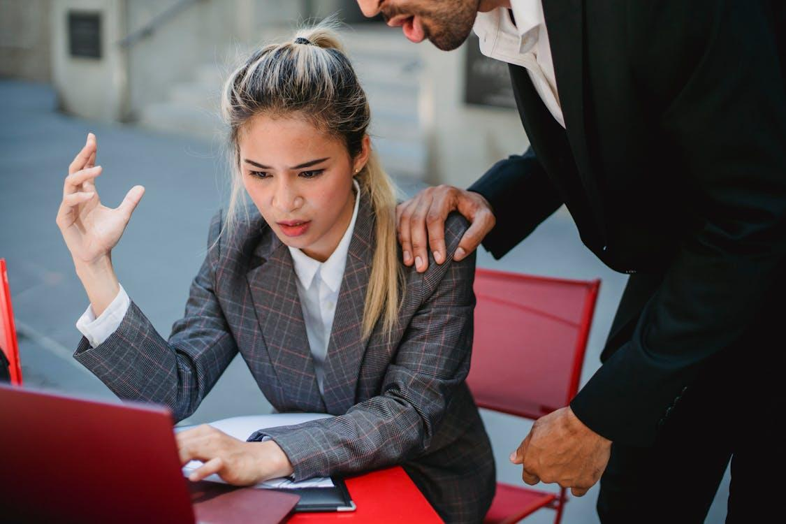 kenson Investments | Retail Meets Digital: Walmart and Amazon Eye Stablecoins in the U.S. A woman in a plaid blazer sitting at a red table looking frustrated at her laptop, while a man stands beside her with his hand on her shoulder.