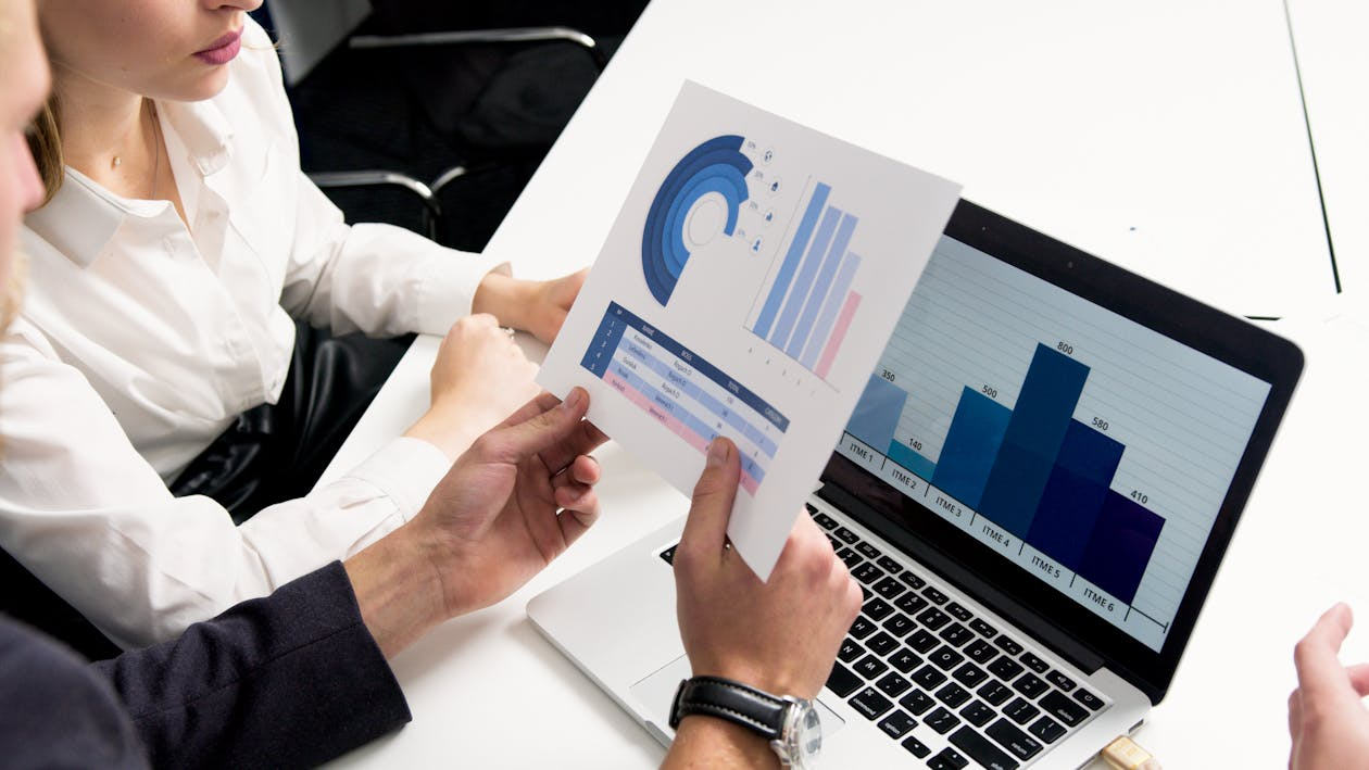 kenson Investments | Is XRP Really Gunning for SWIFT’s Turf? Let’s Talk Numbers Two people reviewing a printed chart with bar graphs and circular data visuals, while a laptop on the table displays a blue bar graph.