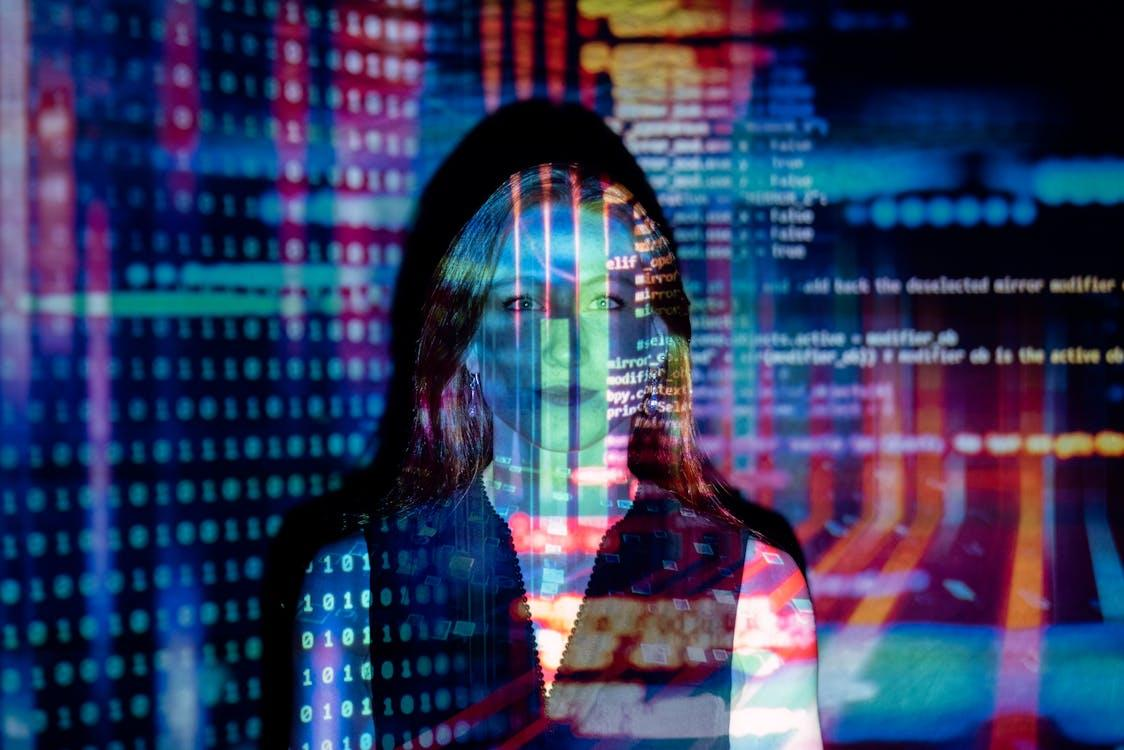 A woman standing in front of a screen with multicolored digital code and binary numbers projected onto her face and clothing.