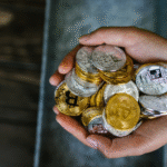 Two hands holding a large pile of silver and gold-colored physical cryptocurrency tokens, including Bitcoin and Ethereum.