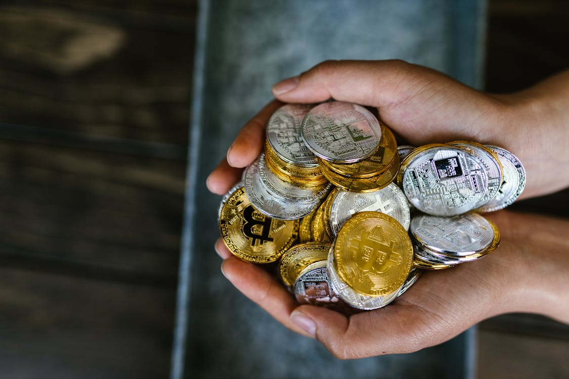 Two hands holding a large pile of silver and gold-colored physical cryptocurrency tokens, including Bitcoin and Ethereum.