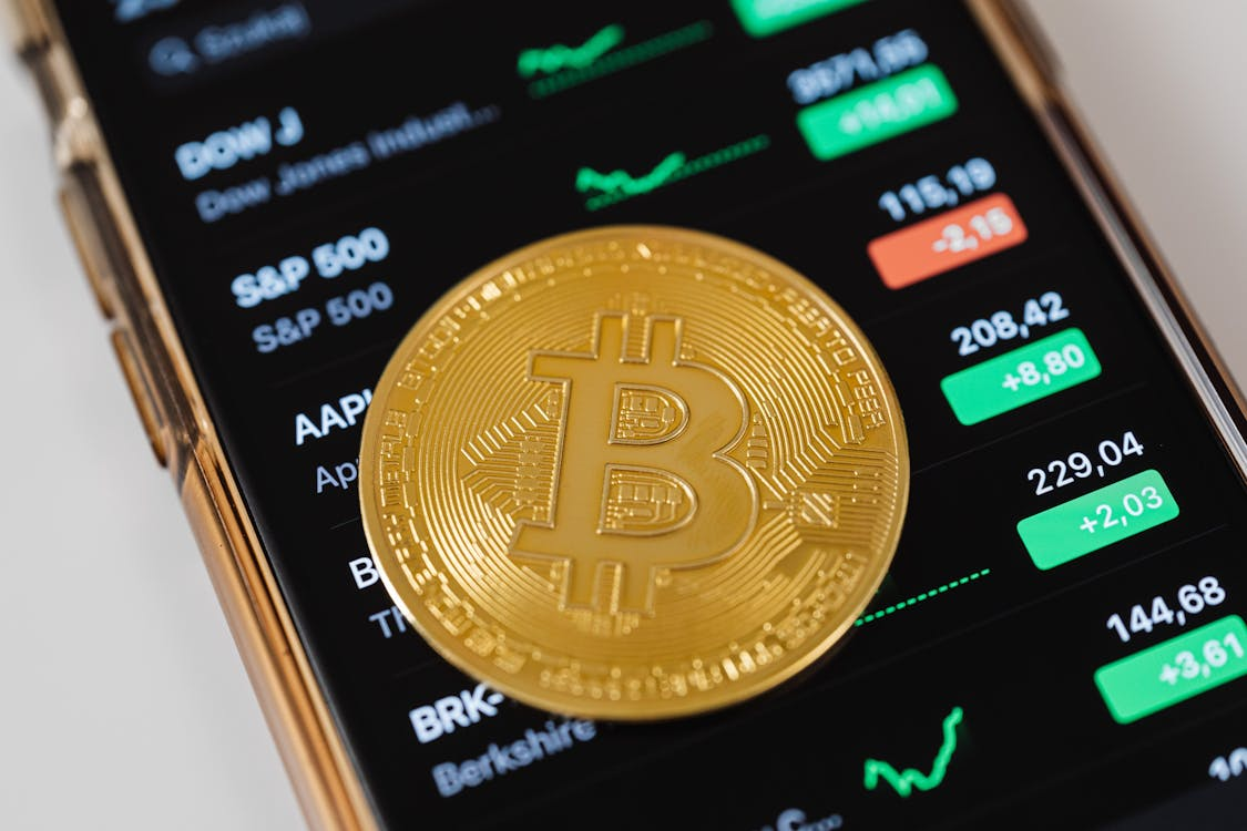 kenson Investments | Whales Are Surfacing: Big ETH Moves Amid Retail Retreat A physical gold-colored Bitcoin placed on top of a smartphone screen displaying a stock market app with various indices and price movements.