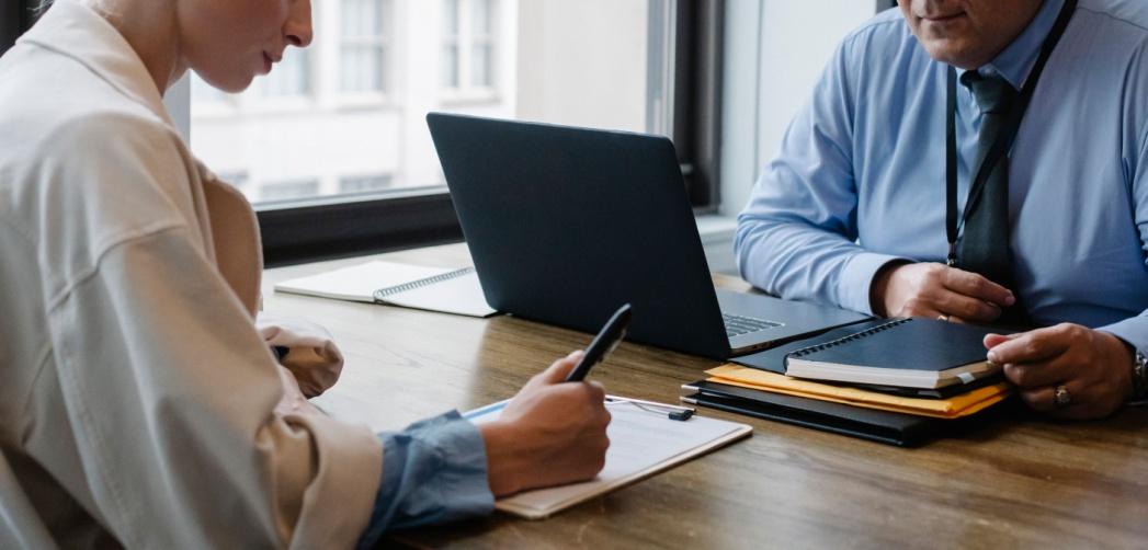 Business professionals reviewing and signing formal documents beside a laptop at an office desk.