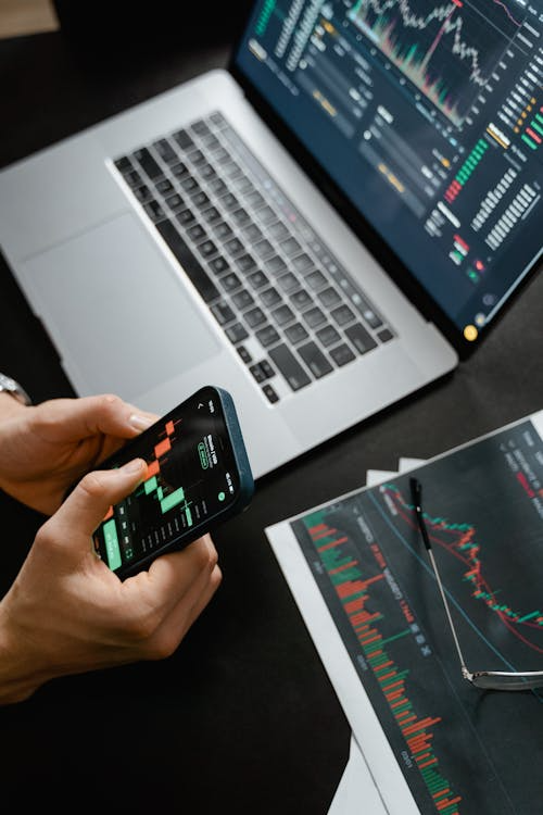 Person holding a smartphone while referencing a laptop displaying charts and graphs, symbolizing real-time digital asset monitoring.