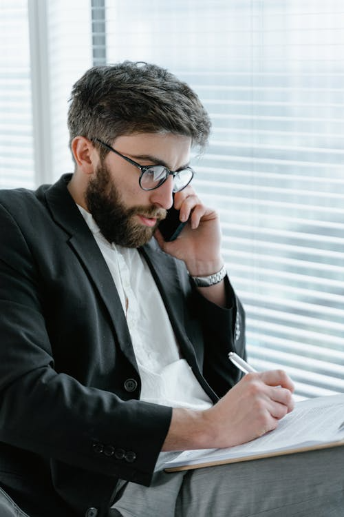 Man in a black suit and glasses, on a call while writing notes, representing active decision-making and process documentation.