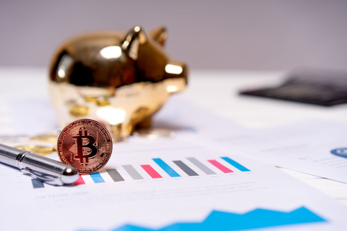 A Bitcoin coin placed near a gold piggy bank and pen on papers displaying financial charts and graphs, symbolizing digital asset management.