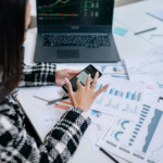 professional analyzing digital asset charts on laptop and mobile device with financial reports on desk