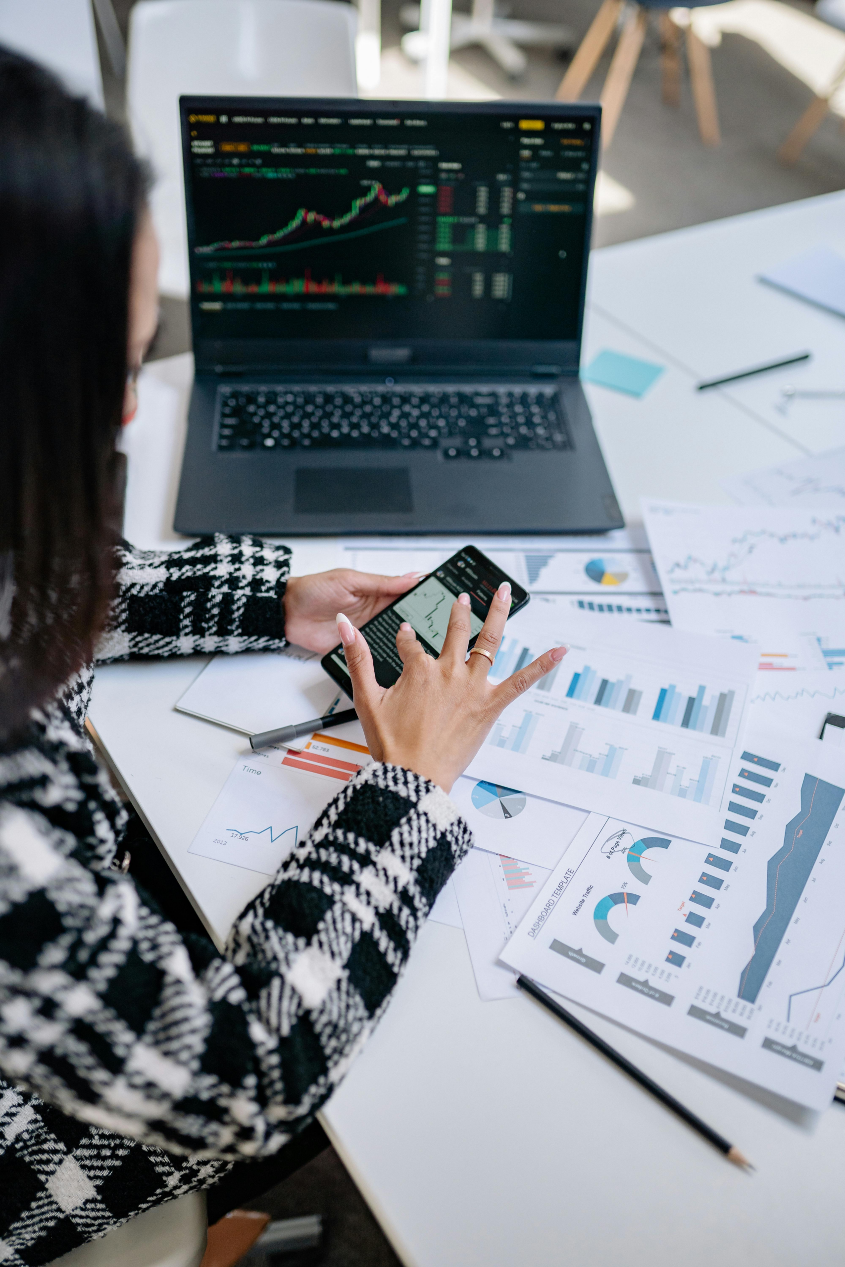 professional analyzing digital asset charts on laptop and mobile device with financial reports on desk