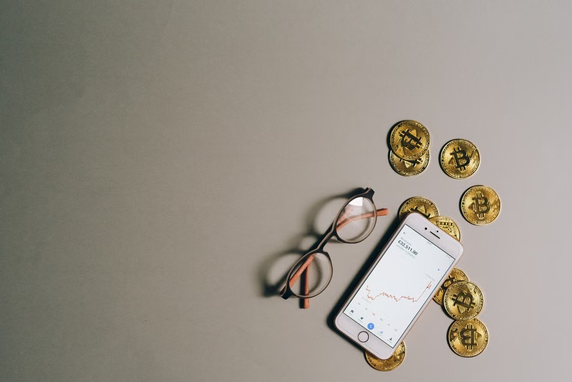 kenson Investments | Proactive Wallet Hardening: Managing UTXO Exposure Before the Quantum Era Smartphone showing data trend chart lying next to eyeglasses and physical Bitcoin tokens.