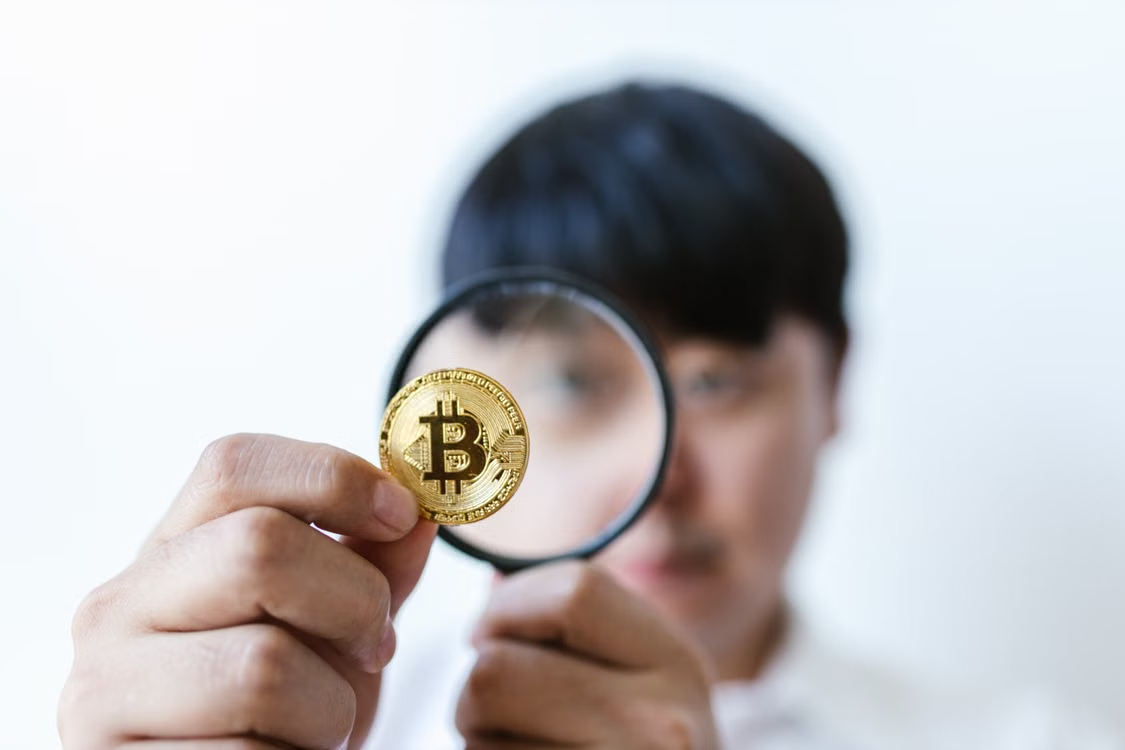 kenson Investments | The Multi-Custodian Imperative Under Regulatory Scrutiny Person inspecting a physical bitcoin token through a magnifying glass closely.