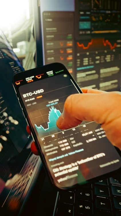 Hand holding a smartphone displaying live cryptocurrency market trading charts.