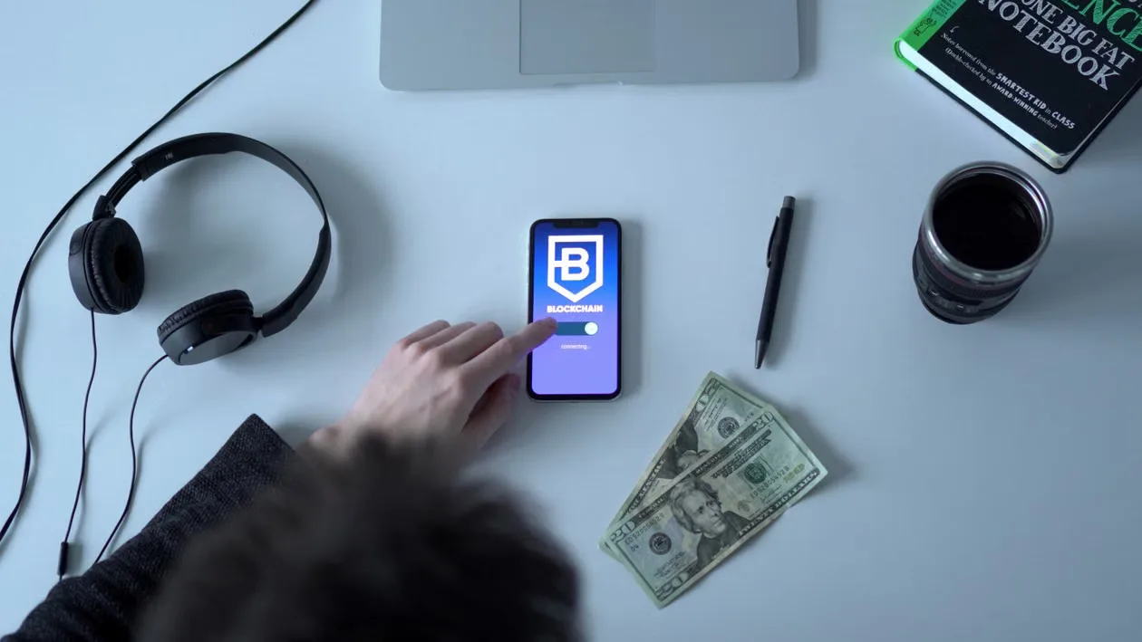 Overhead view of blockchain app on smartphone with cash, headphones, and notebook.