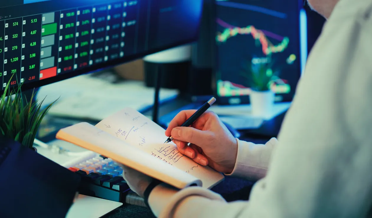 Person analyzing financial data on screens, making notes